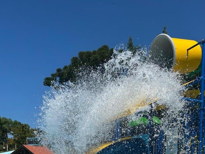Balde gigante amarelo virando água sobre o brinquedo infantil, com árvores e céu azul ao fundo
